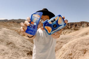 Person showcasing sunscreen and camera in a sunny desert setting, promoting sun protection.