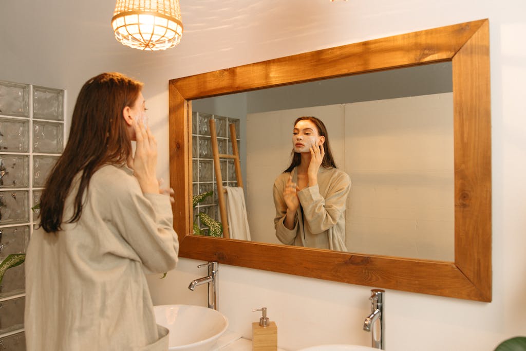 Woman applying skincare cream in front of a mirror in a cozy bathroom setting.